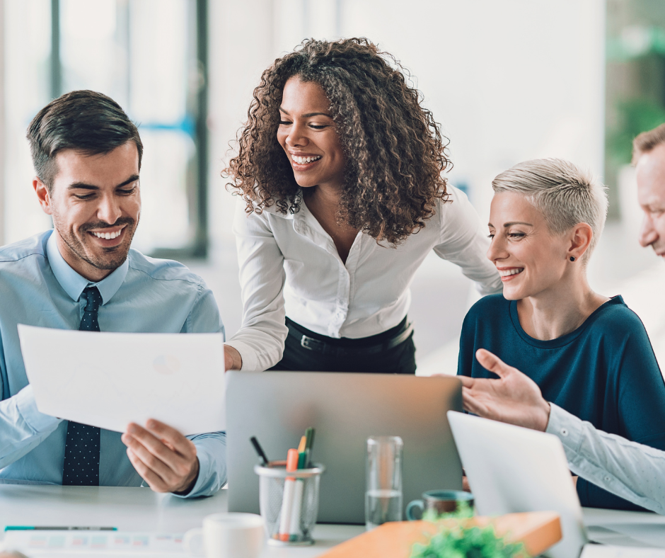 Four colleagues smiling and discussing documents around a laptop in a bright office.