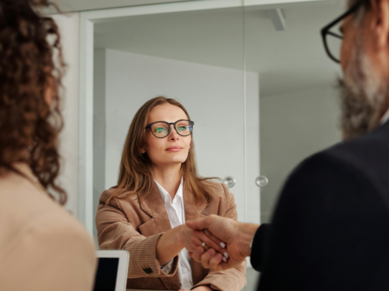 Woman in glasses and brown blazer shaking hands with a man in a suit during a meeting.