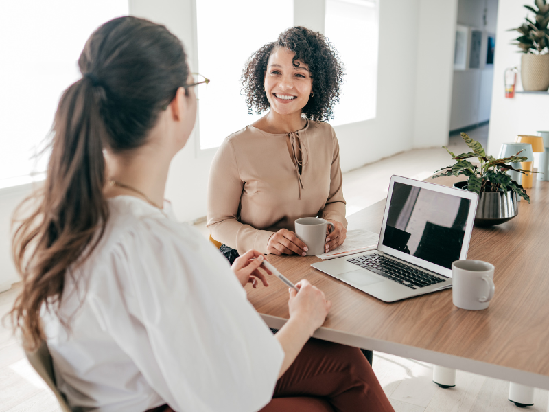 Two women sitting at a table in a bright office, smiling and talking with a laptop and coffee mugs on the table.