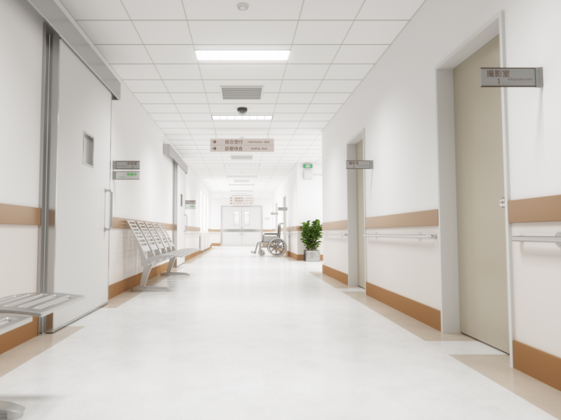 Bright hospital corridor with white walls, beige accents, metal benches, a wheelchair near double doors, and signs in Japanese indicating rooms and directions.