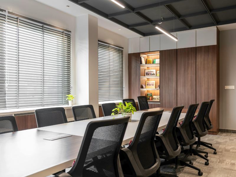Modern conference room with a long white table, black mesh office chairs, large windows with blinds, and wooden cabinets with shelves.