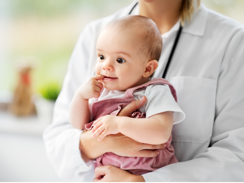 Baby wearing pink overalls with finger in mouth being held by a person in a white medical coat.