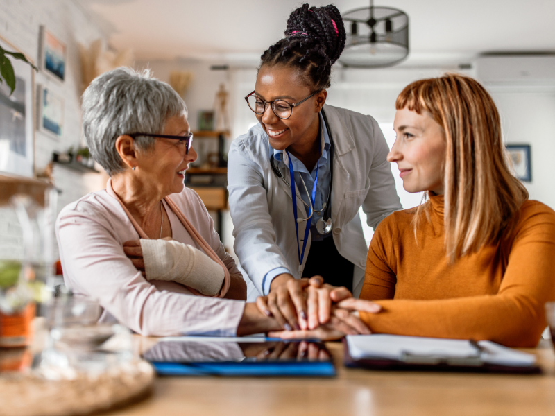 Smiling female doctor placing her hand on the hands of two women, one elderly with an arm in a cast and one younger, in a supportive and friendly gesture at a table with documents and a tablet.