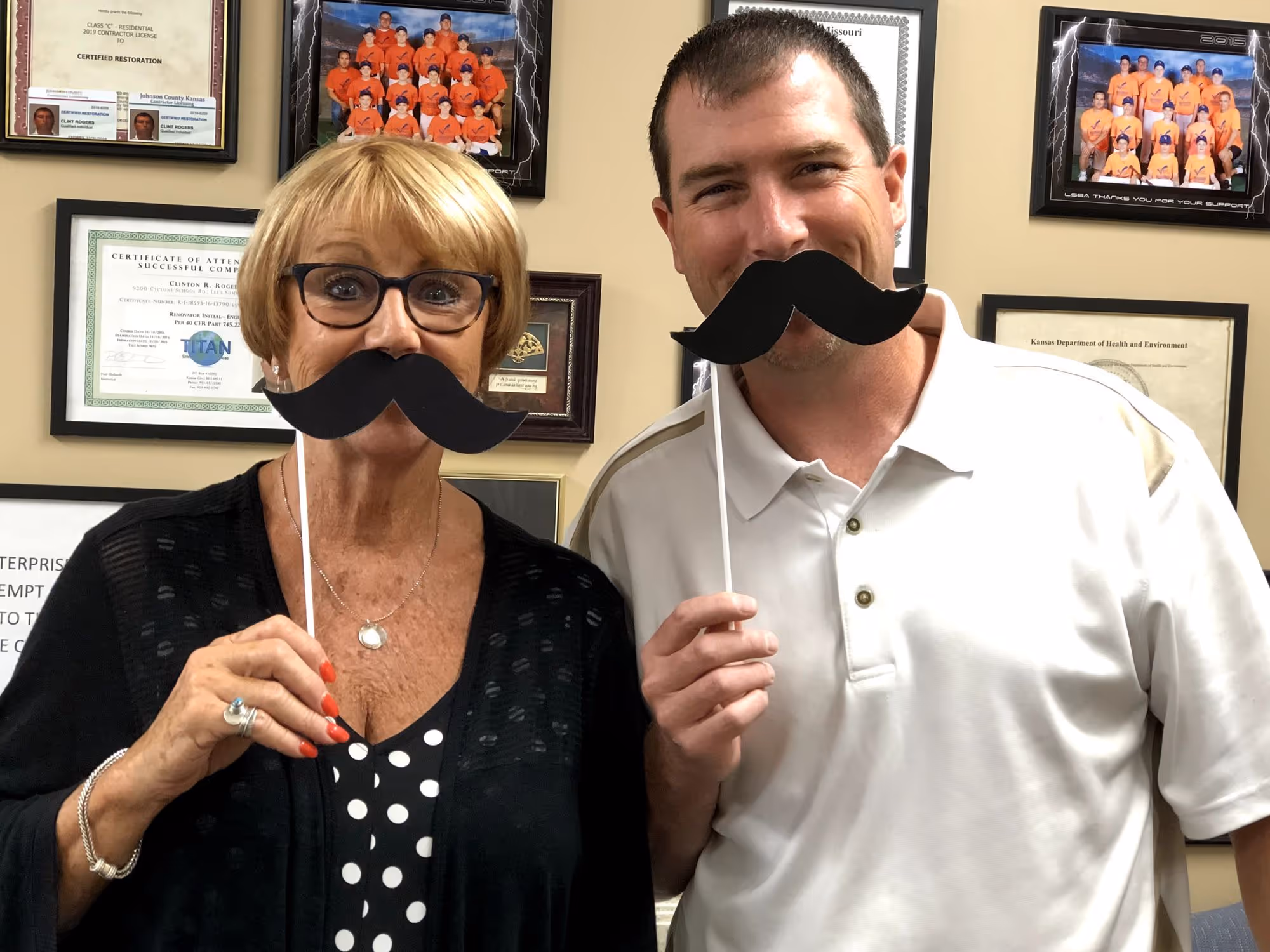 A woman and a man holding black paper mustaches on sticks in front of their faces while smiling, with certificates and framed photos on the wall behind them.