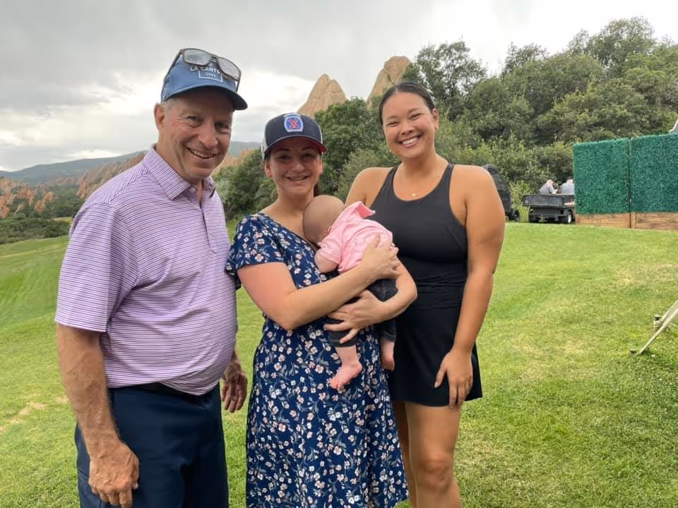 Three adults smiling outdoors on grass with trees and rocky hills in the background; one woman holds a baby dressed in pink.