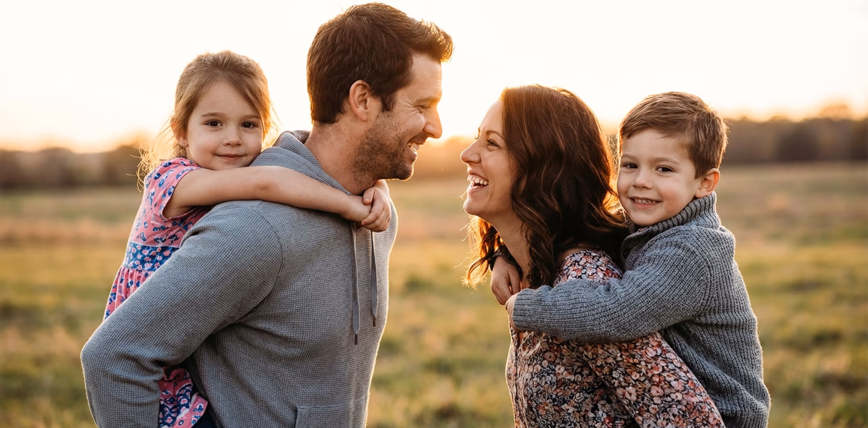 Smiling young family outdoors at sunset with parents giving piggyback rides to their children.