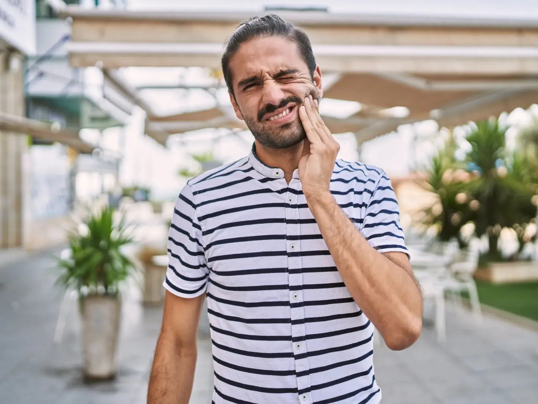 Man with man bun wearing a striped shirt, touching his cheek in pain outdoors.