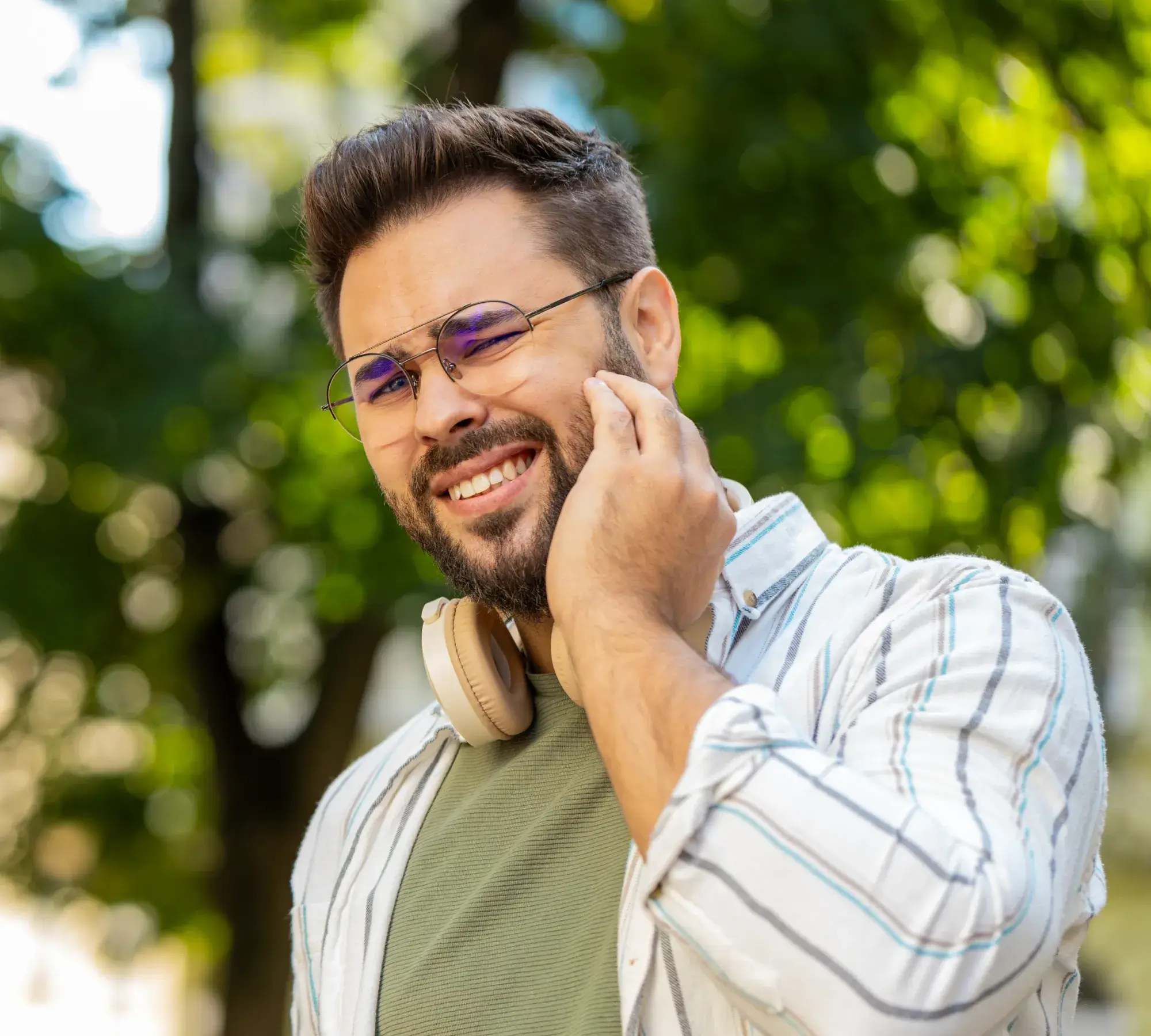 Man with glasses and headphones wincing in pain while touching his jaw outside with blurred green background.