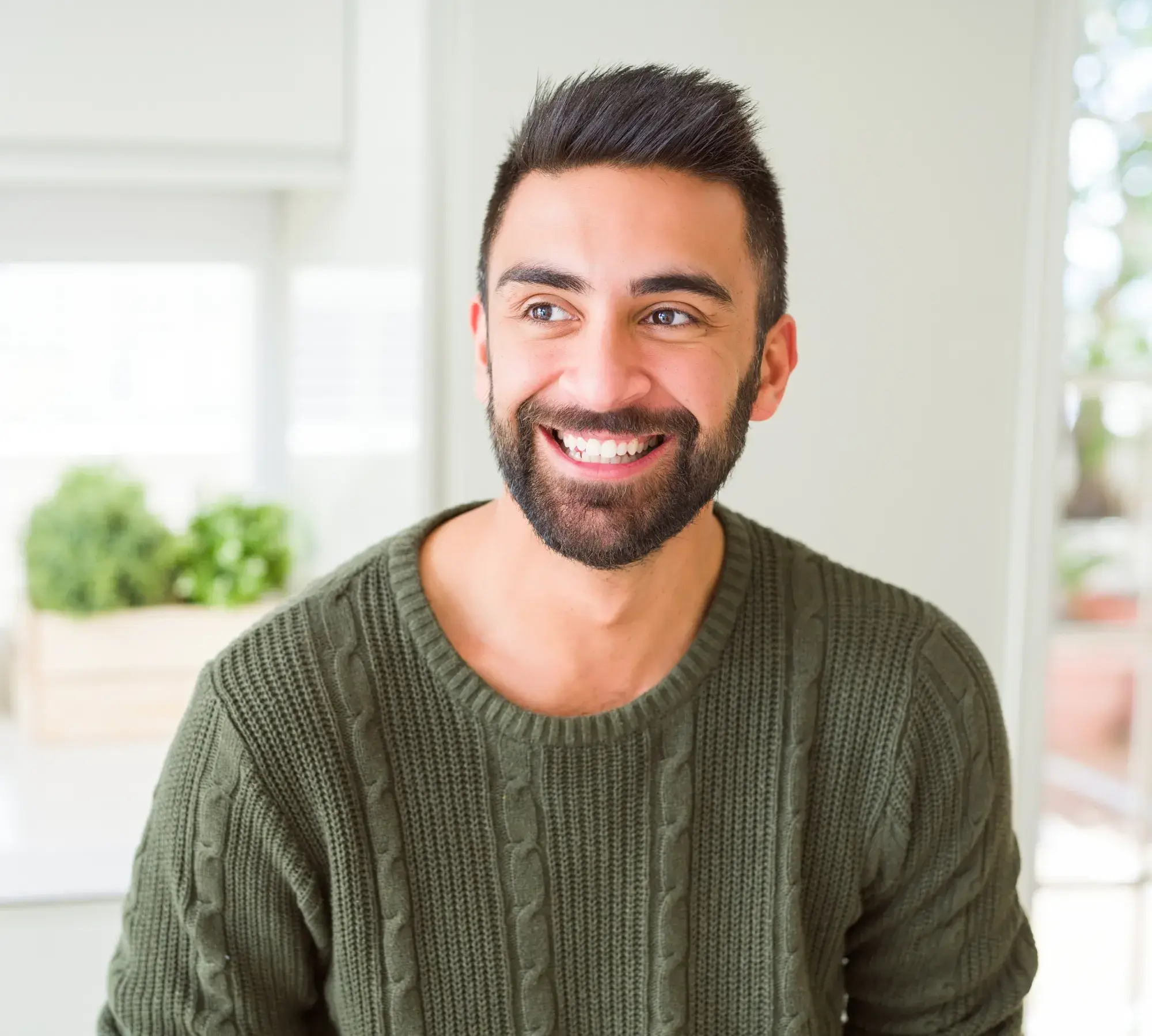 Smiling man with beard and short dark hair wearing a green cable-knit sweater in a bright room.