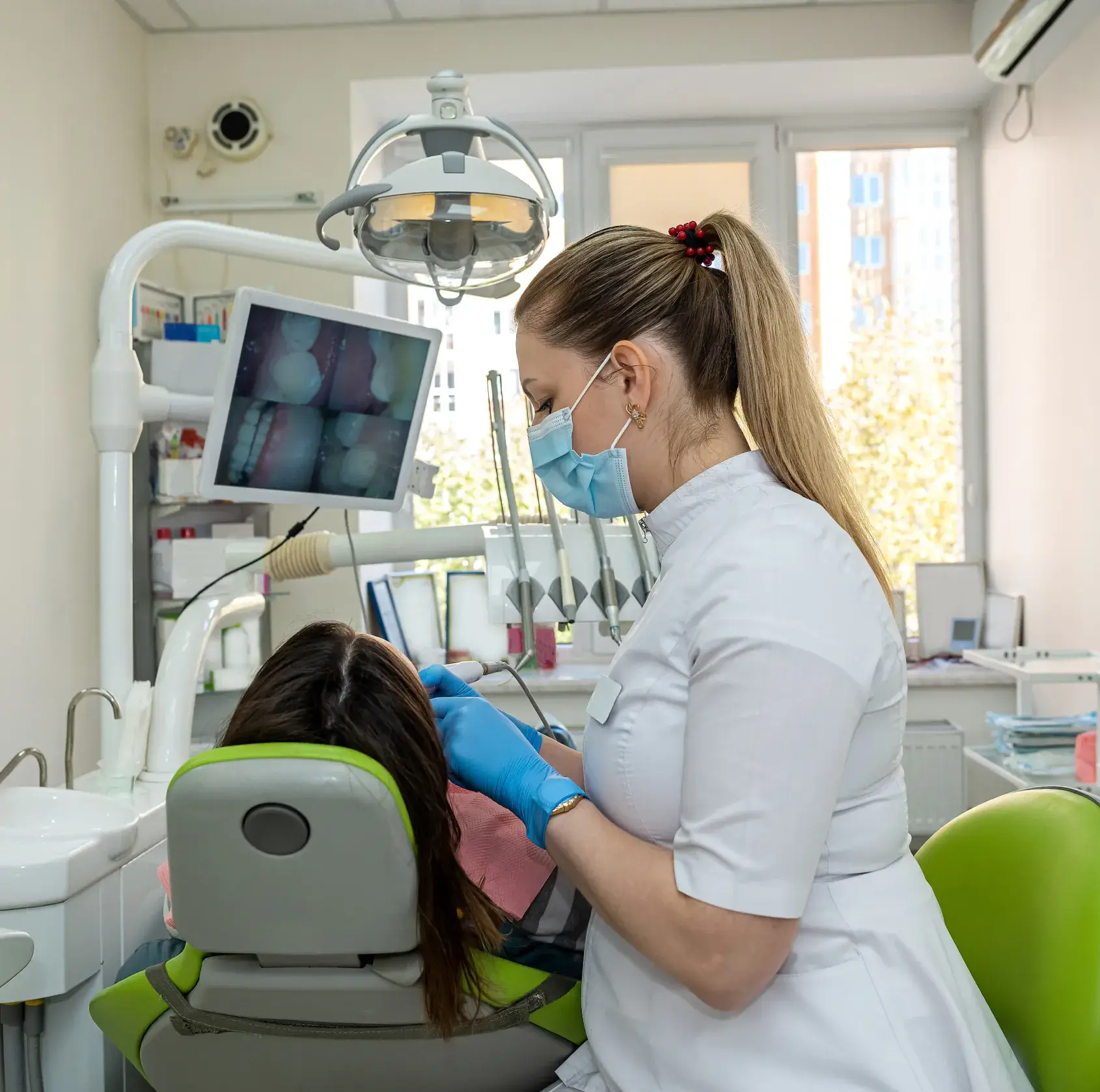 Dentist wearing a mask and gloves examining a patient's teeth in a dental clinic with dental equipment and an X-ray image on the monitor.