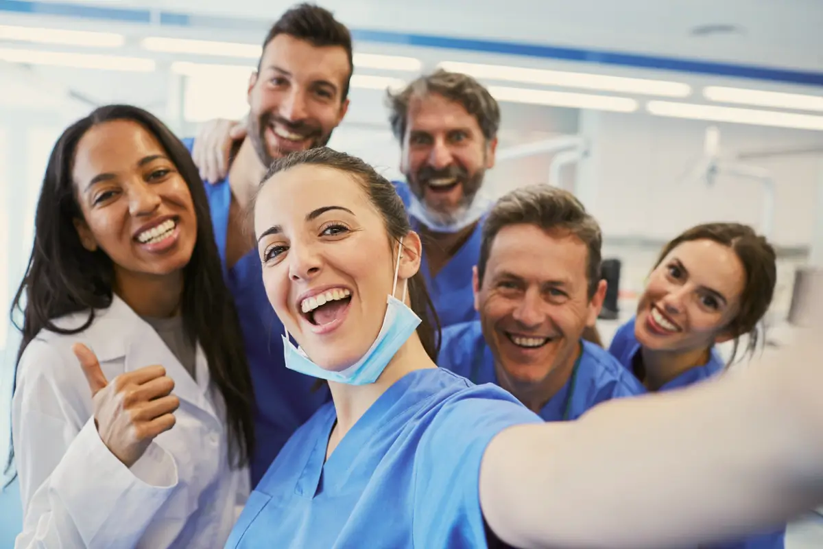 Group of six diverse healthcare professionals smiling and taking a cheerful selfie in a medical setting.