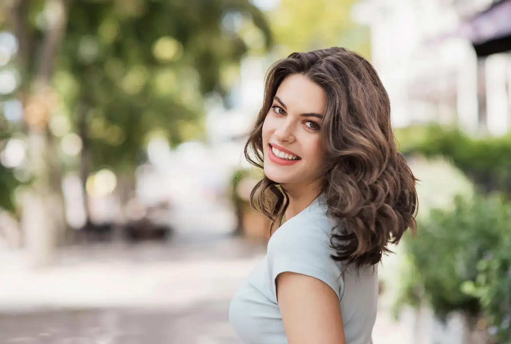 Smiling young woman with wavy brown hair in a light blue shirt outdoors with blurred green foliage background.