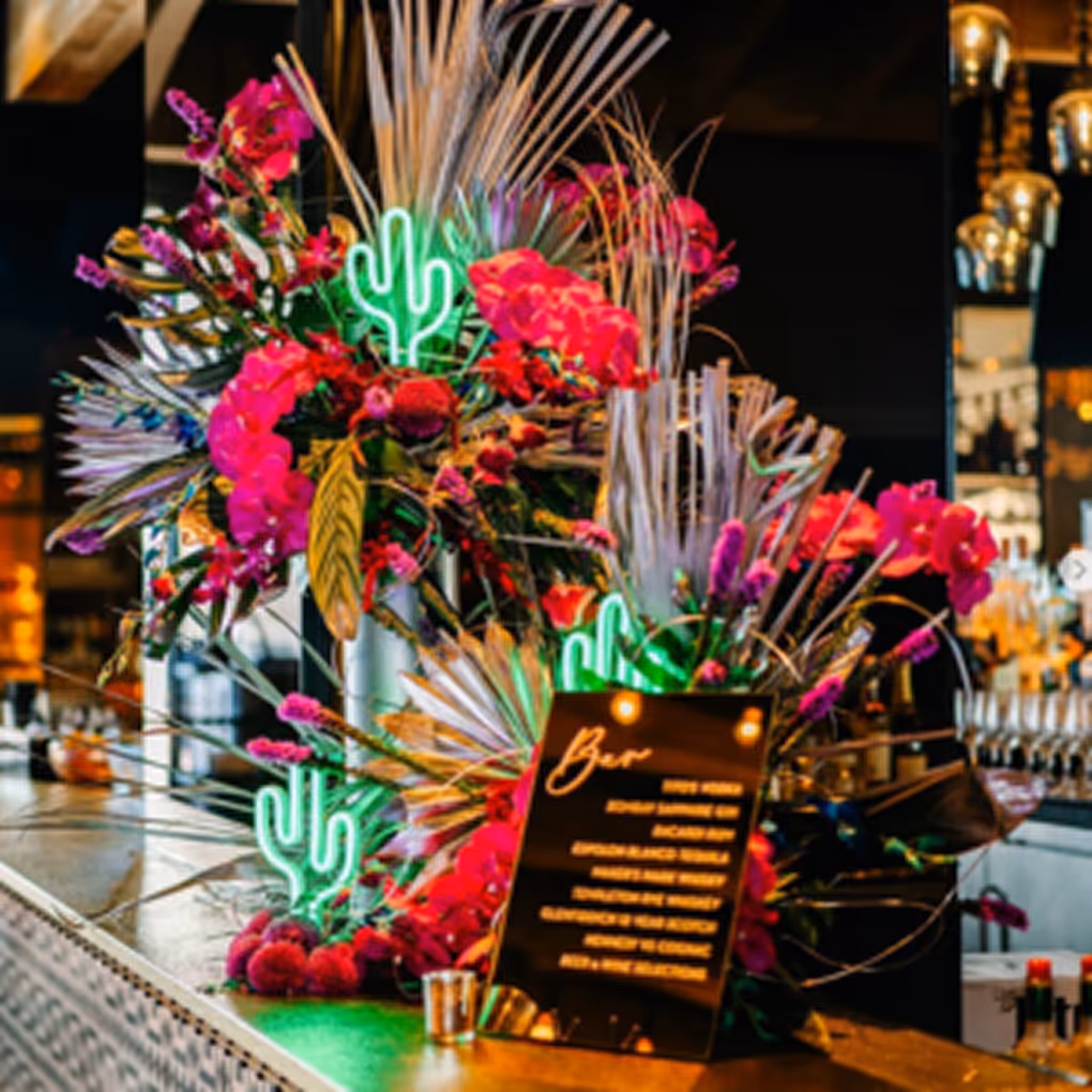Bar countertop decorated with vibrant pink flowers, dried palm leaves, neon green cactus lights, and a menu board listing drinks.