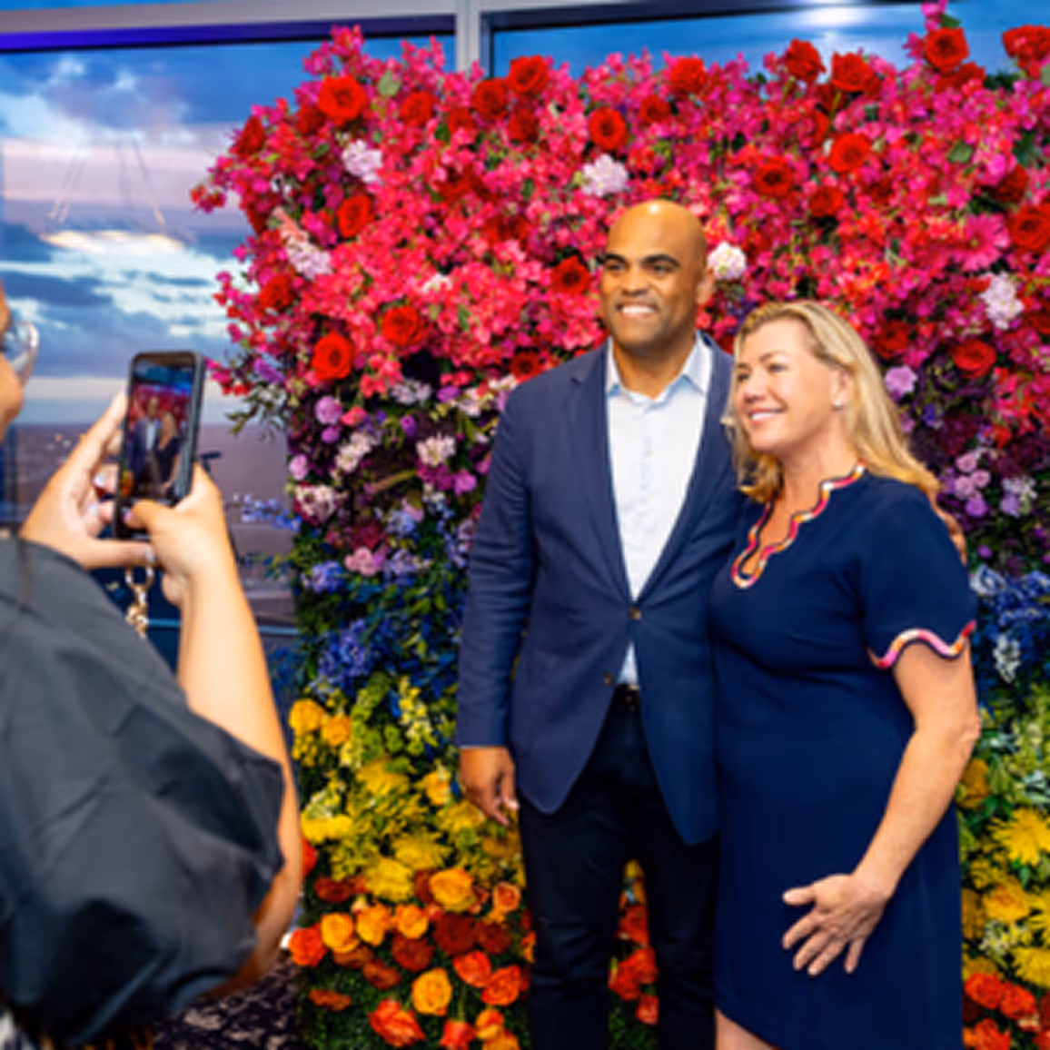 A man in a blue suit and a woman in a navy dress pose for a photo in front of a colorful flower wall while someone takes their picture with a smartphone.