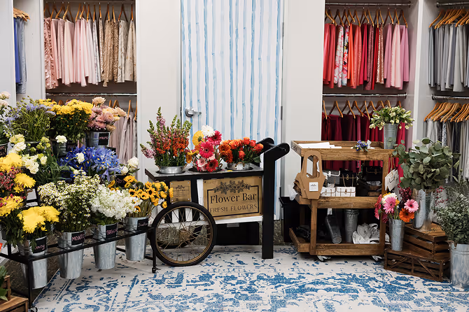 Indoor flower shop with colorful fresh flowers arranged in metal buckets, a flower cart labeled 'Flower Bar Fresh Flowers,' and clothing racks with neatly hung garments in the background.