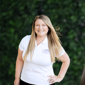 Smiling woman with long blonde hair wearing a white polo shirt standing outdoors with green trees in the background.