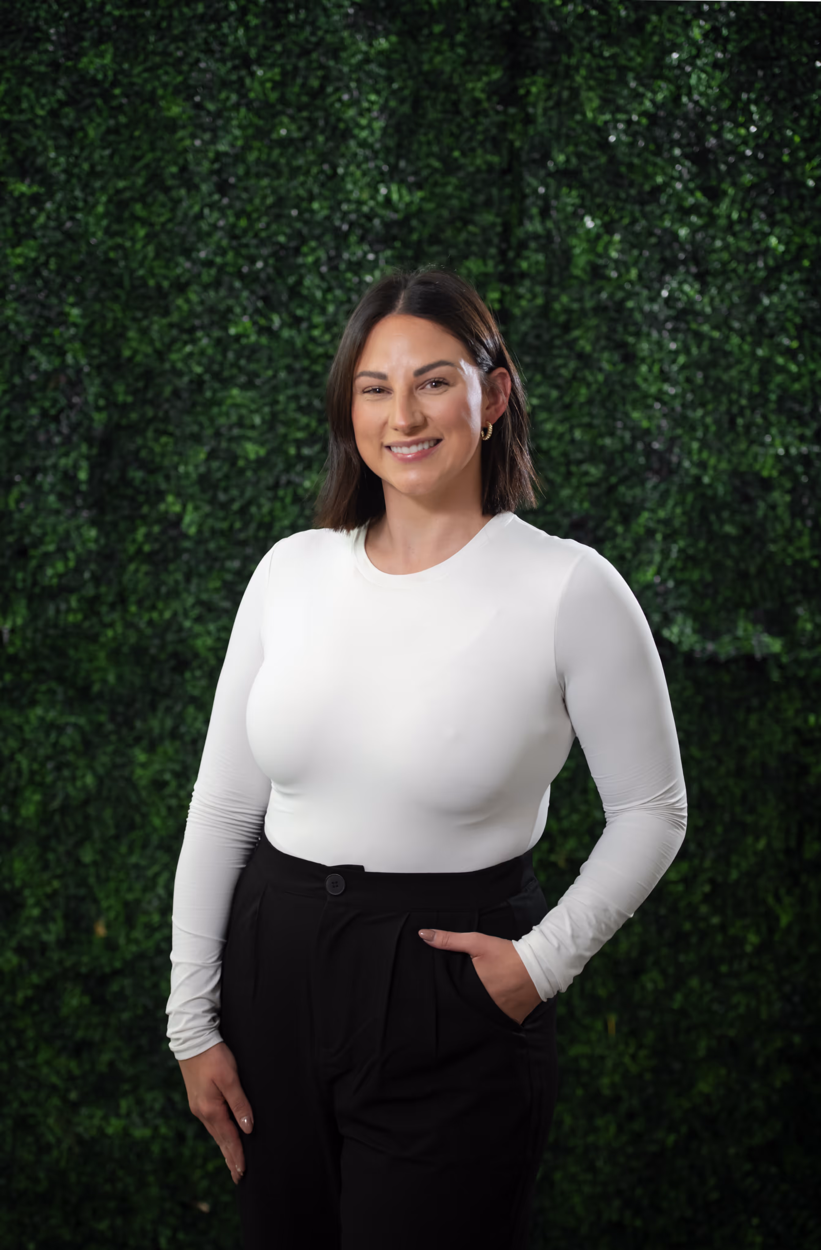 Smiling woman with shoulder-length dark hair wearing a white long-sleeve top and black pants standing in front of a green leafy background.