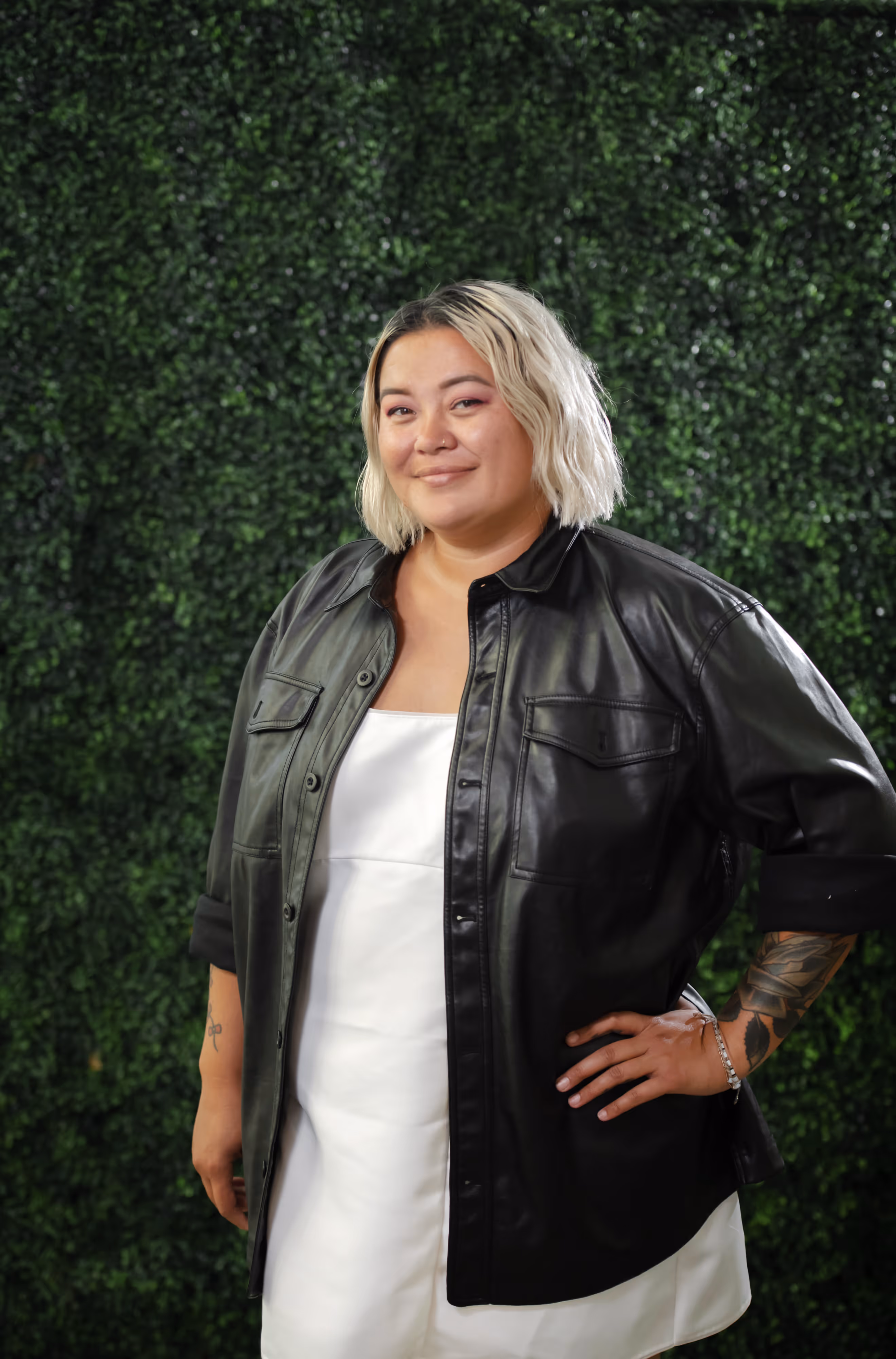 Person with short bleached hair wearing a white dress and black leather jacket standing with hand on hip against a green leafy backdrop.