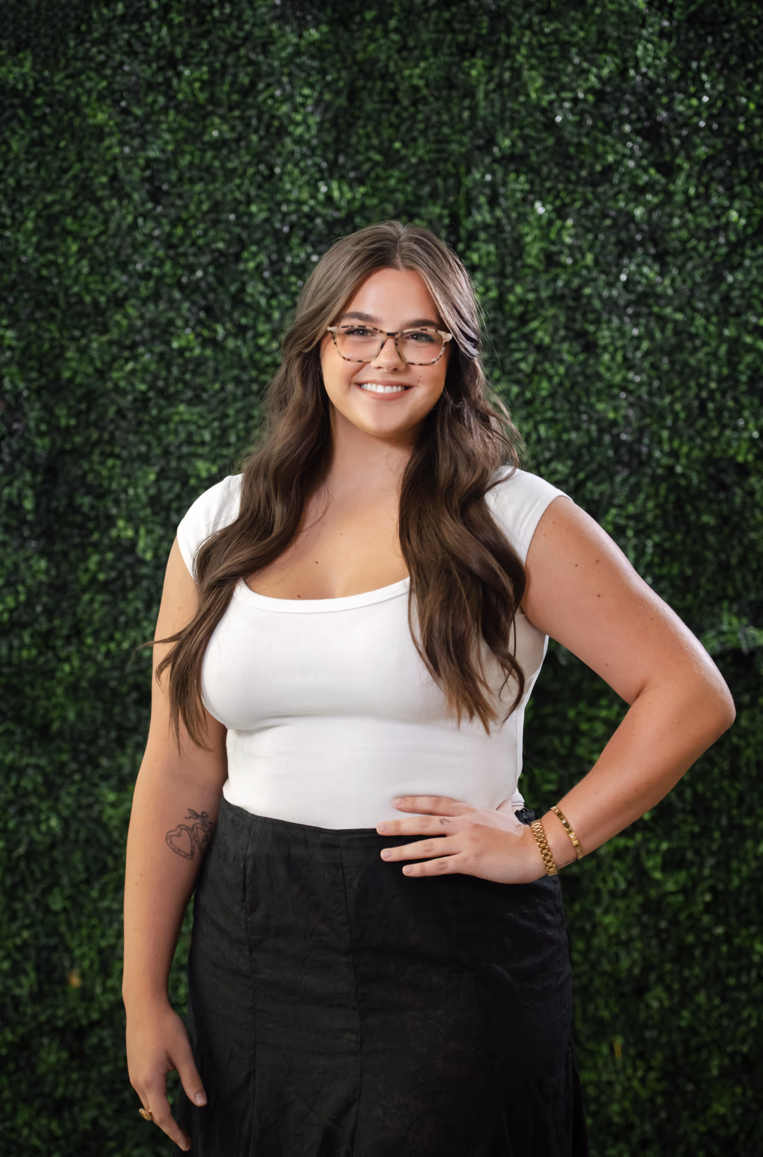 Smiling woman with long brown hair wearing glasses, a white top, and black skirt standing against a green leafy background.