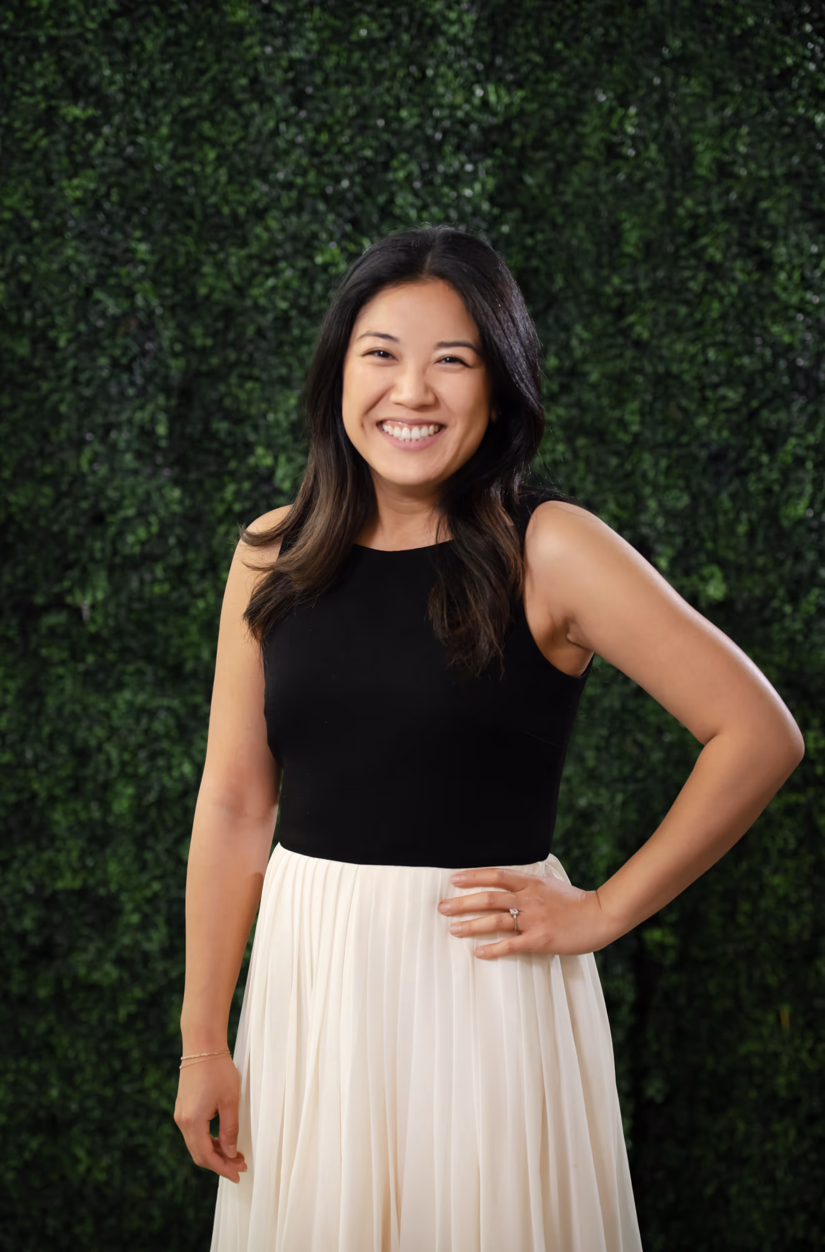 Smiling woman with long dark hair wearing a black sleeveless top and white pleated skirt standing against a green leafy background.