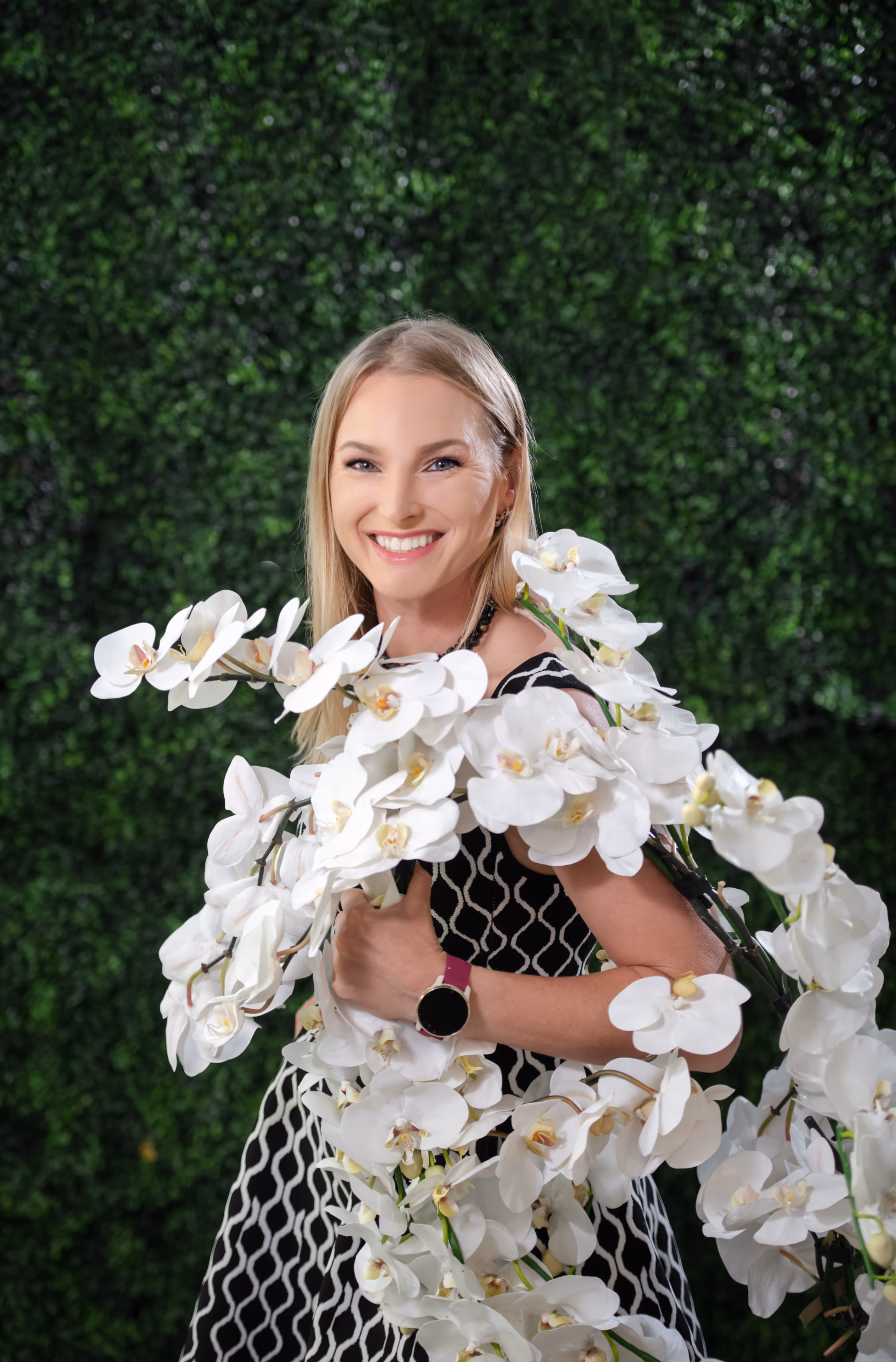 Smiling blonde woman in a black and white patterned dress holding a large bouquet of white orchids against a green leafy background.