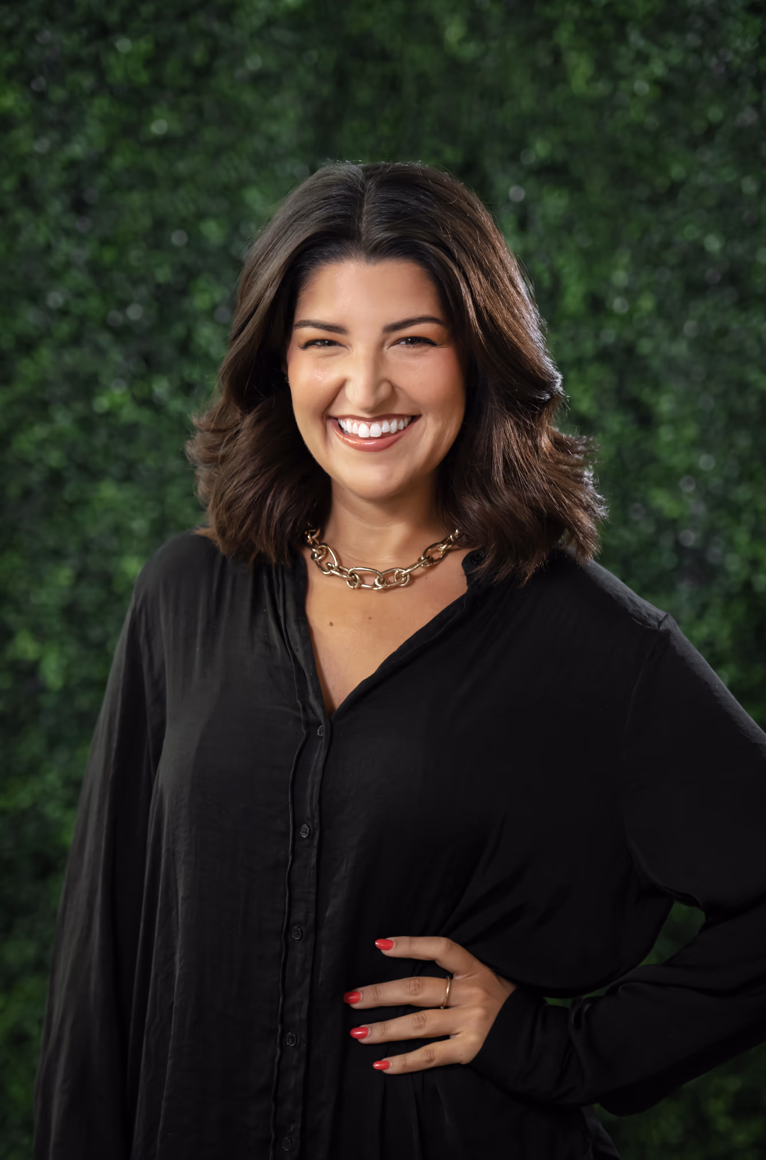 Smiling woman with dark wavy hair wearing a black buttoned shirt and gold chain necklace, standing in front of a green leafy background.