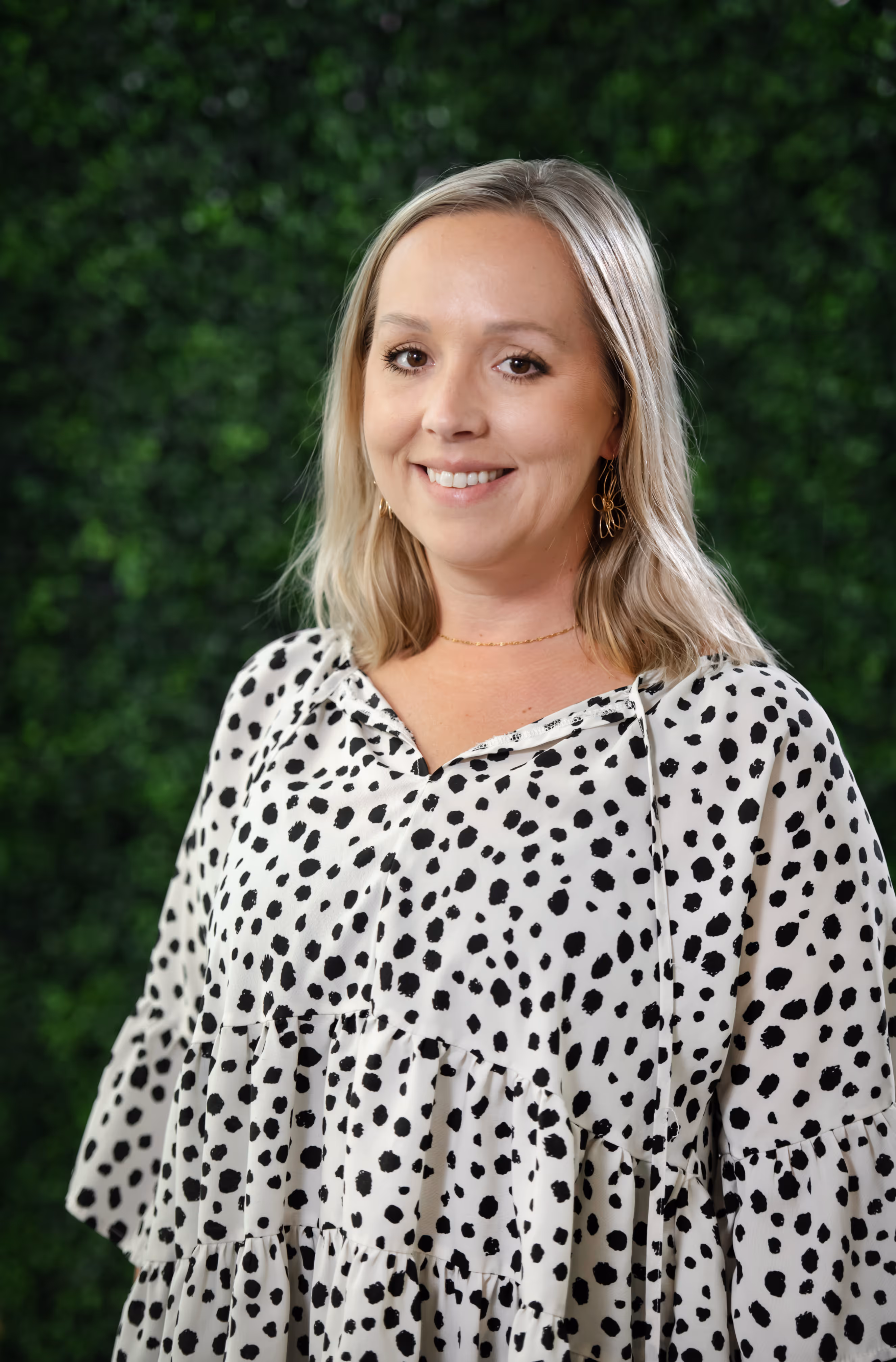 Smiling woman with blonde hair wearing a white dress with black polka dots, standing against a green leafy background.