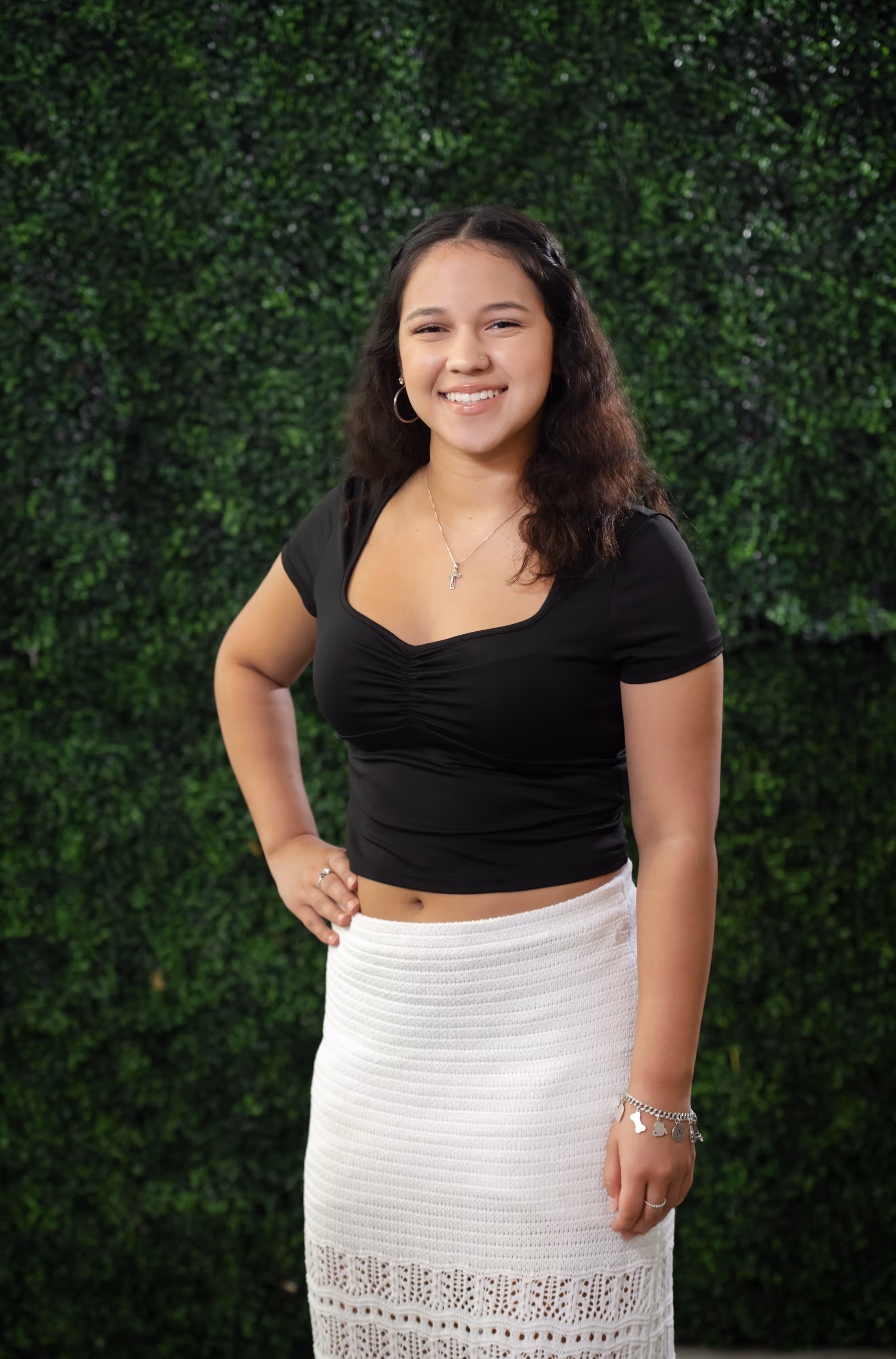 Smiling young woman with curly hair wearing a black top and white skirt standing in front of a green leafy background.