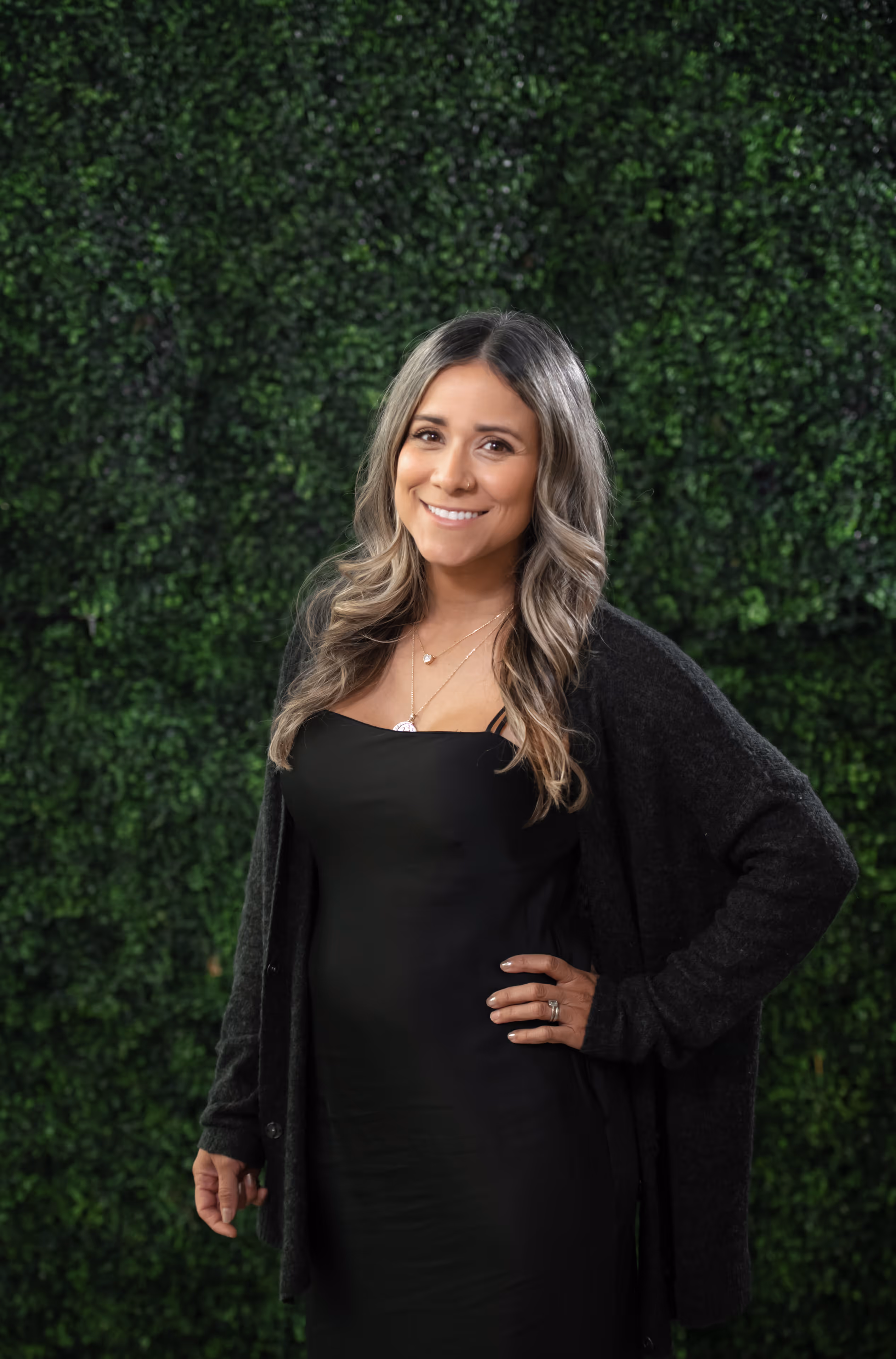 Smiling woman with long, wavy hair wearing a black dress and cardigan, standing in front of a green leafy background.
