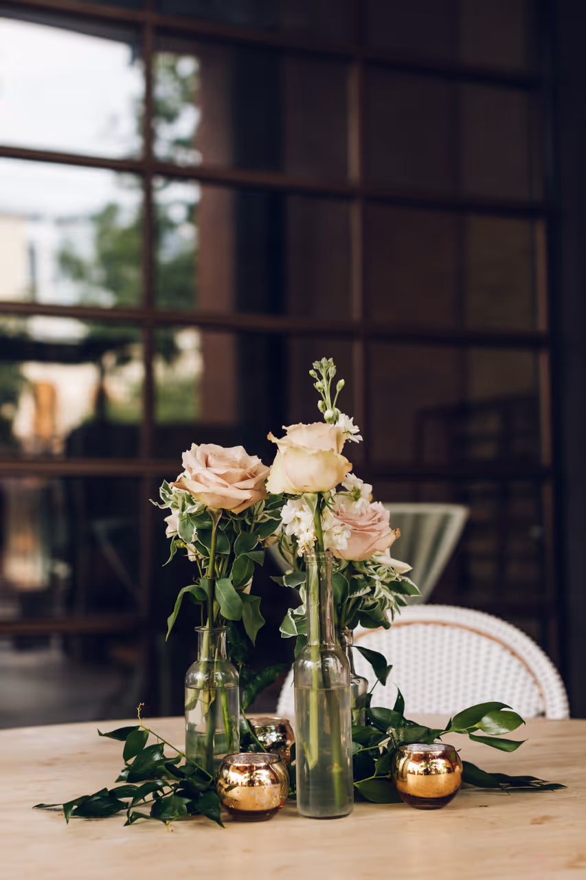 Three glass bottles with pale pink and white roses and greenery arranged on a wooden table with three small gold candle holders.