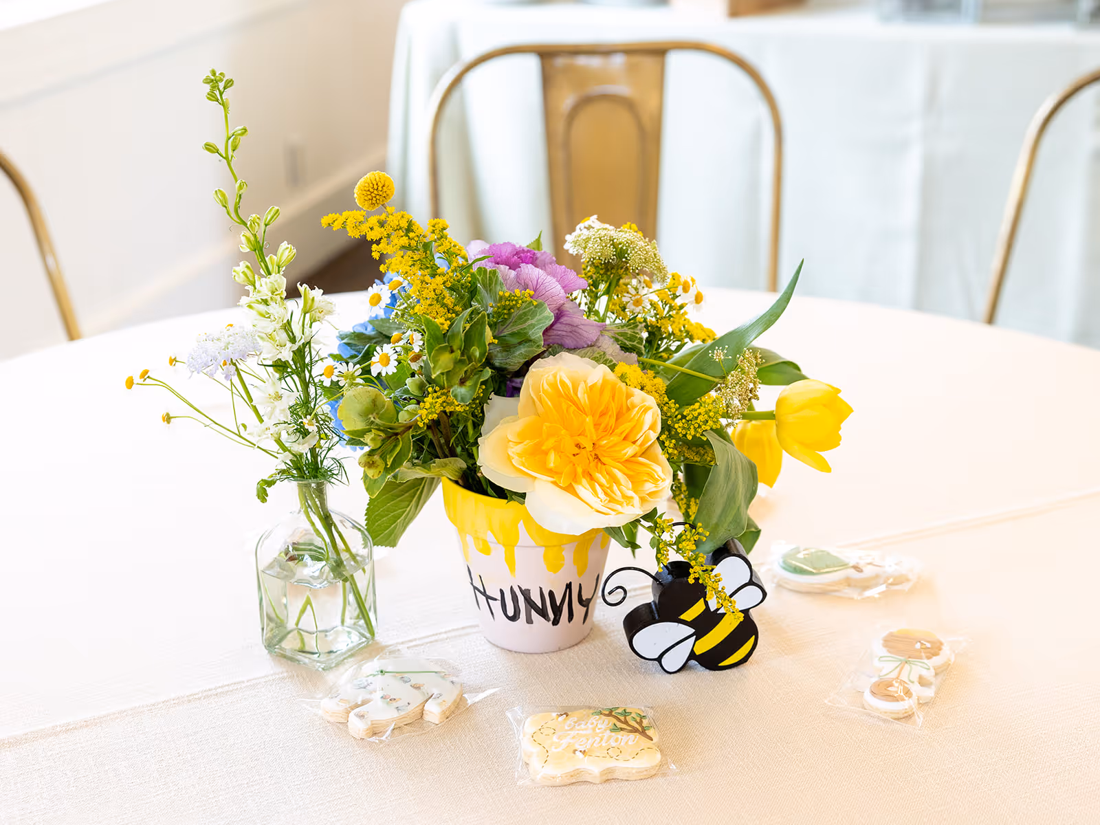 Colorful flower arrangement in a pot labeled 'HUNNY' with a decorative bee and small vases with flowers on a white tablecloth.