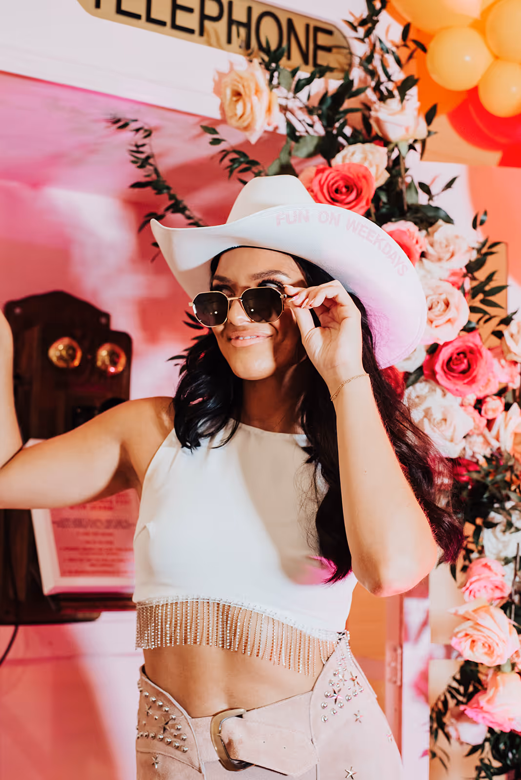 Woman wearing a white cowboy hat with text, sunglasses, a white cropped top with fringe, and high-waisted pants, posing in front of a vintage telephone and floral backdrop.