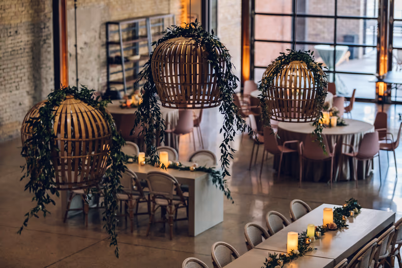 Indoor event space with wooden lanterns wrapped in greenery hanging over tables decorated with candles and green foliage.