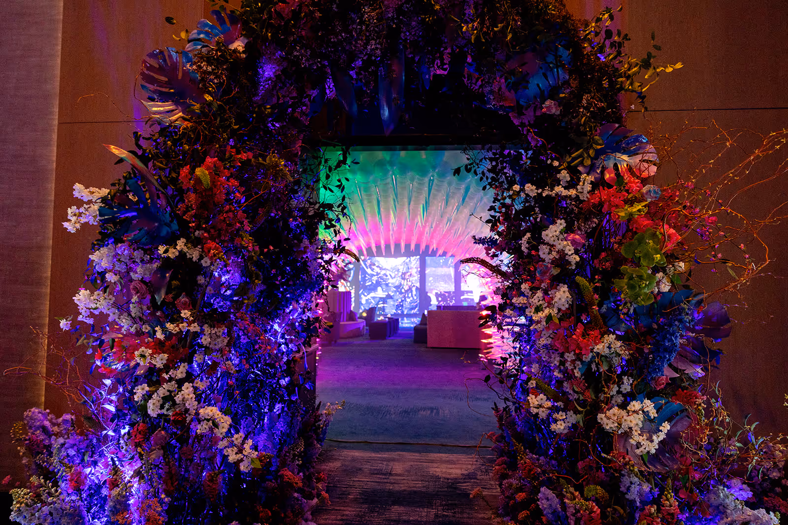 Archway decorated with colorful flowers and illuminated with purple and blue lights, leading to a room with neon green, pink, and purple lighting.