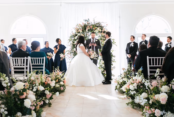Bride and groom standing at the altar during a wedding ceremony with an officiant and guests seated along flower-lined aisle.