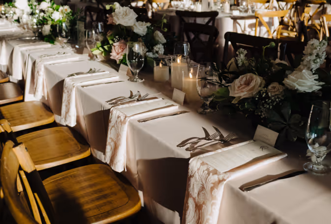 Elegant wedding reception table with white tablecloth, floral centerpieces, candles, and wooden chairs.