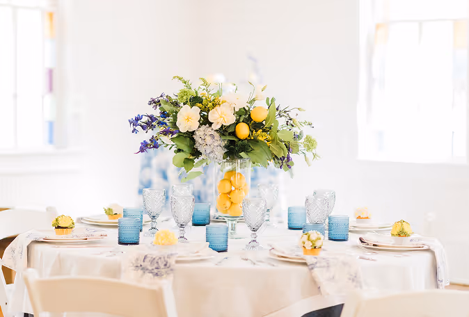 Round table set with blue glassware, white plates, and a centerpiece of mixed flowers and lemons in a glass vase.