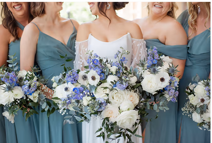 Bride in white dress and bridesmaids in teal dresses holding bouquets of white and purple flowers.