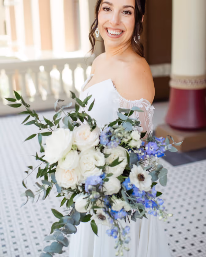 Smiling bride in off-shoulder white dress holding a bouquet of white and blue flowers with green leaves.