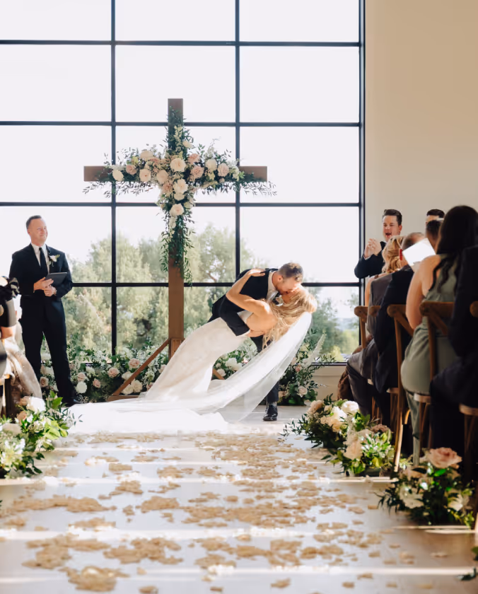 Bride and groom sharing a romantic dip kiss at the altar during their wedding ceremony in a bright chapel with floral decorations and a large cross.