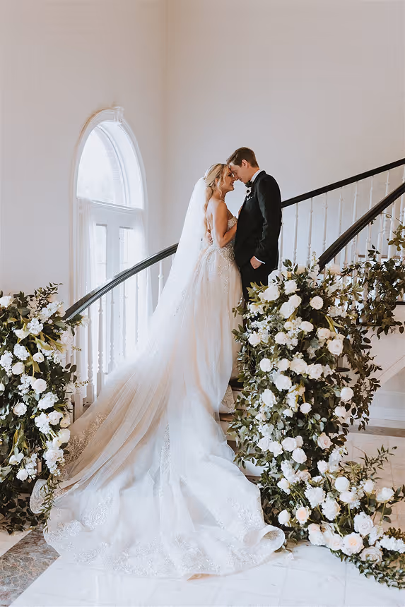 Bride and groom standing close on a decorated staircase with white flowers and greenery, bride wearing a long white gown with a veil.