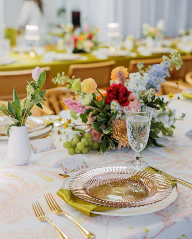 Elegantly set dining table with gold cutlery, crystal glass, pink decorative plate, green napkin, and vibrant floral centerpiece.