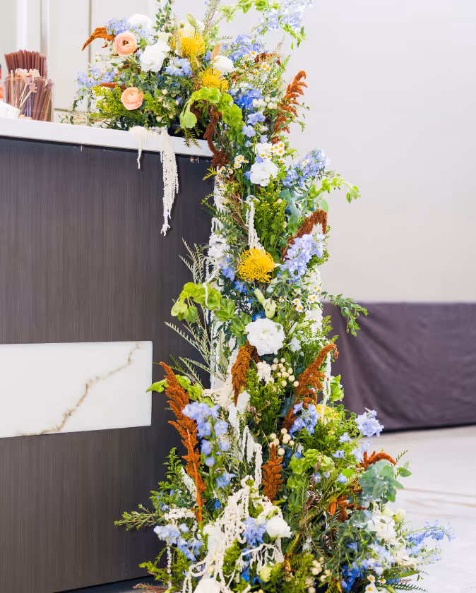 Tall floral arrangement with white, blue, yellow, and orange flowers cascading beside a dark wood and white marble counter.