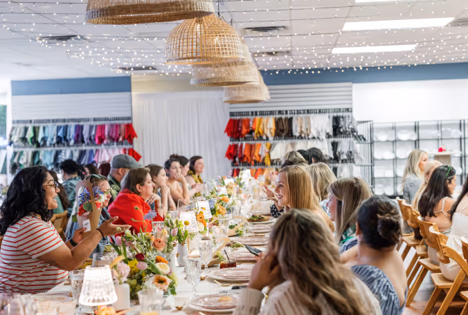 People sitting around a long table decorated with flowers and table settings during a social event under woven pendant lights and string lights.