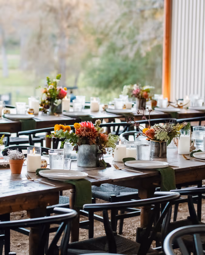 Rustic wooden tables set for an event with plates, glasses, green napkins, candles, and floral centerpieces.