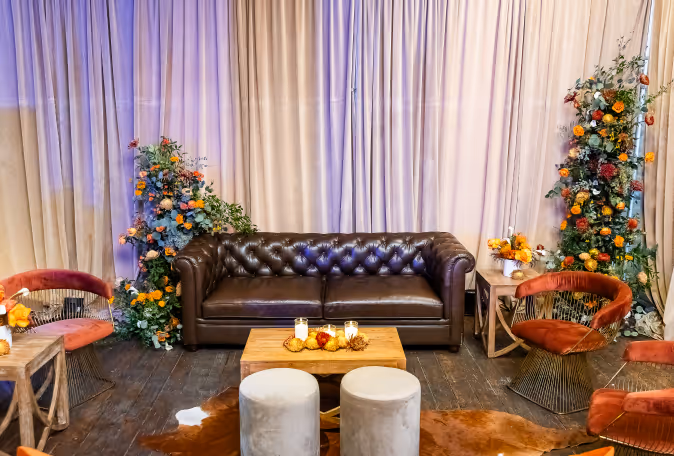 Cozy seating area with a dark brown leather tufted sofa, surrounded by two floral arrangements and rust-colored velvet chairs, centered around a wooden coffee table with candles and decorative items.