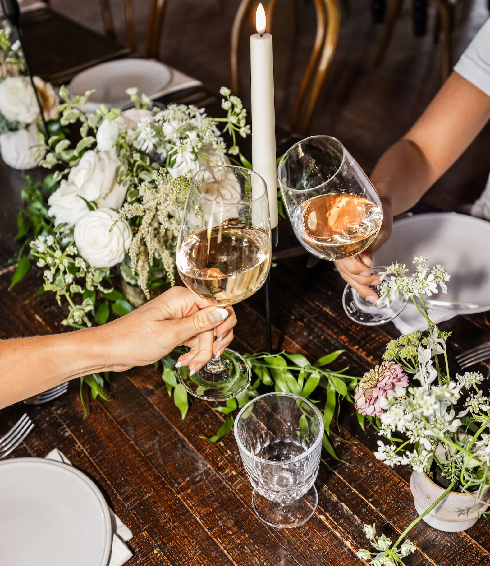 Two people clinking wine glasses over a wooden table decorated with white flowers and a lit white candle.