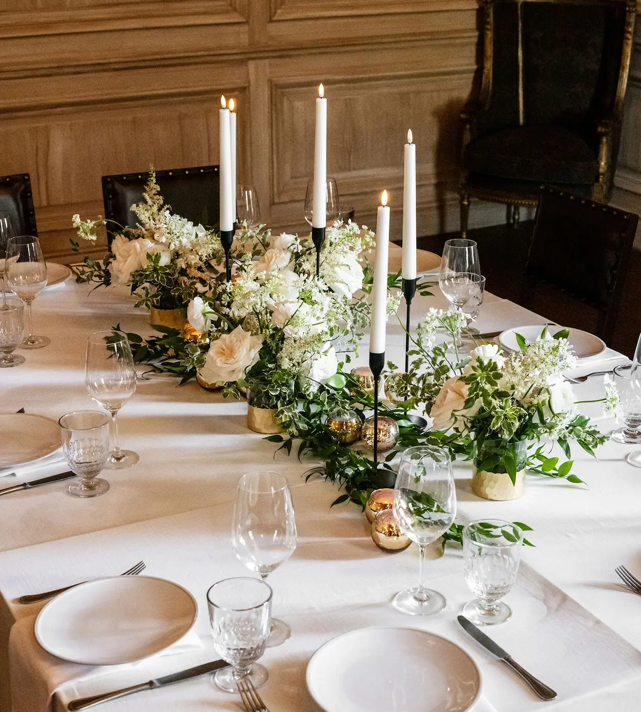 Elegant dining table set with white plates, glassware, and floral centerpiece with white flowers and lit candles.