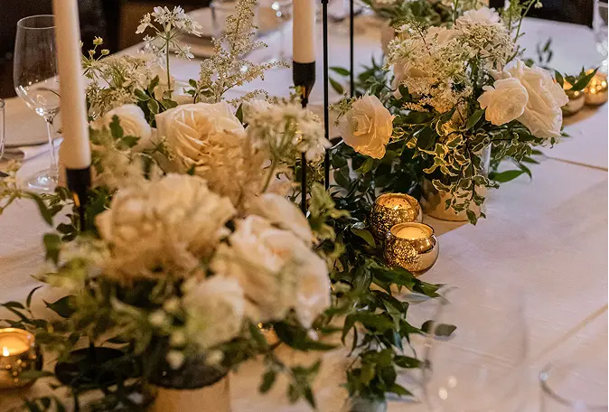 Table centerpiece with white roses, greenery, tall white candles, and small gold candle holders on a white tablecloth.