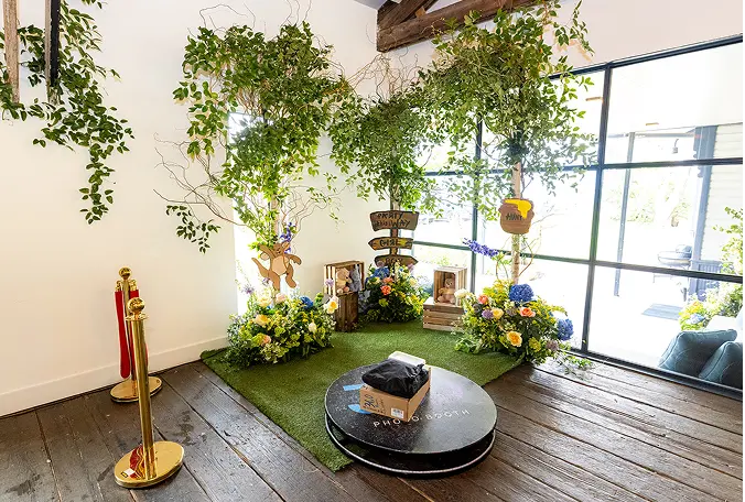 Indoor decorative setup with two small trees surrounded by colorful flowers, cartoon character cutouts, and wooden signs on artificial grass near large windows.